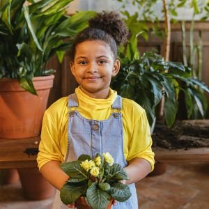 Young girl in overalls holding a plant, surrounded by greenery indoors.